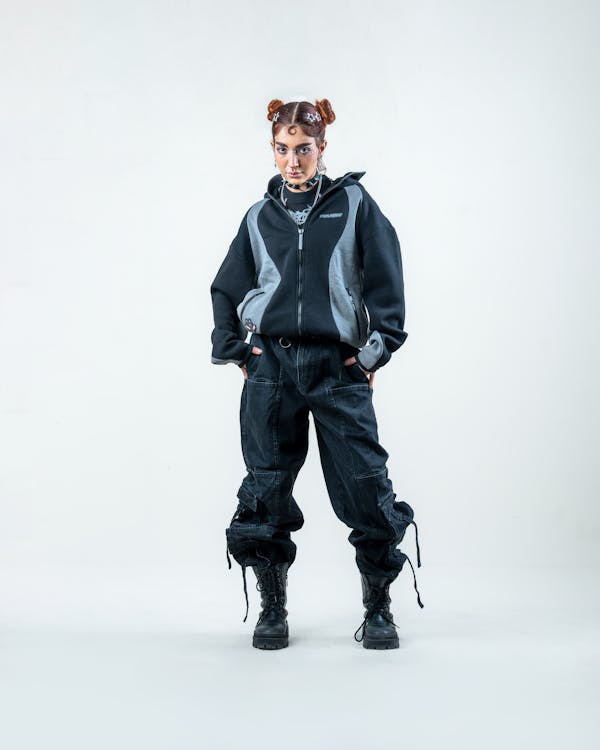 a young woman with red hair styled in space buns is standing against a white background, wearing a black and grey zipup hoodie, dark cargo pants, and chunky black boots. her pose is slightly defiant and confident, hands in pockets. her accessories include necklaces. the overall style is grunge and urban streetwear. the image is a fashion photograph, likely for a clothing brand's advertising.