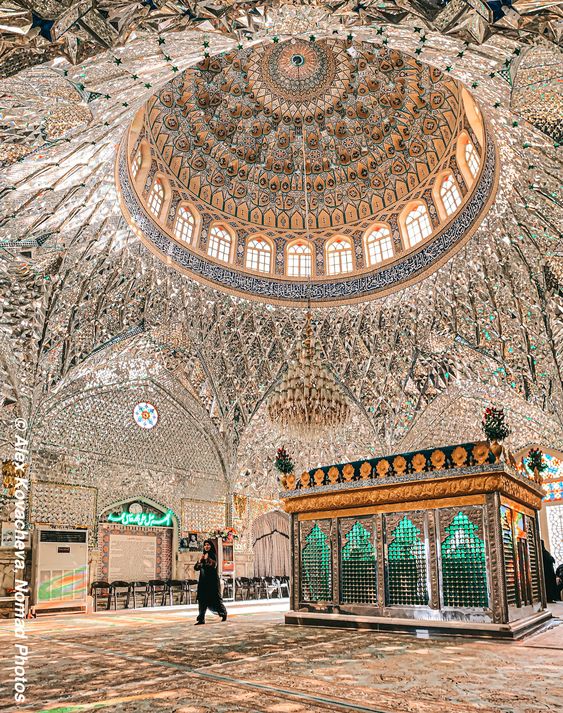this is an interior view of a mosque, possibly a shrine, with a high, mirrored dome and a tomb surrounded by a green, cagelike structure. a woman dressed in black is walking across the open floor. the space is lit by natural light coming through the windows around the base of the dome. the interior is richly decorated with intricate patterns and mirror work.