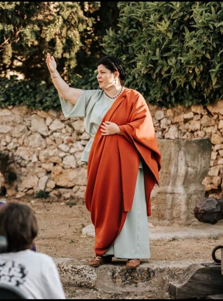 a woman dressed in a romanstyle tunic and orange toga stands before a stone wall, possibly at an archaeological site. she is gesturing with her right hand, seemingly addressing an audience (partially visible in the lower left corner). the setting suggests a historical reenactment or educational presentation about roman history and culture.  the overall mood is one of historical immersion and learning.
