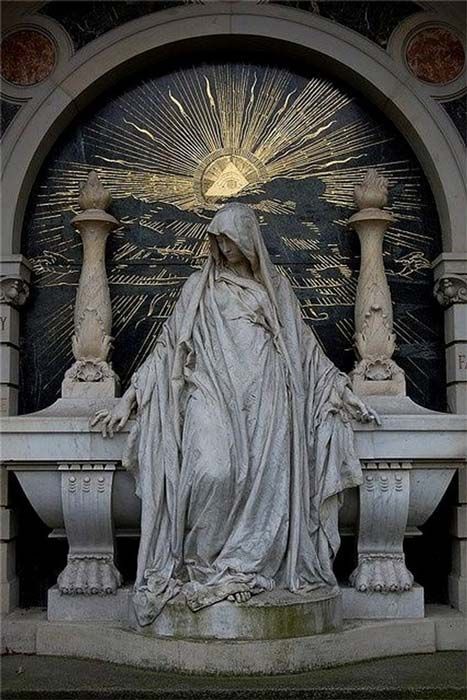 a marble statue of a veiled woman stands in front of a memorial with an archway. behind the statue, a black backdrop is emblazoned with a golden allseeing eye surrounded by rays of light. the woman is draped in heavy robes and appears to be in a state of mourning. the monument is set in what seems to be a cemetery.