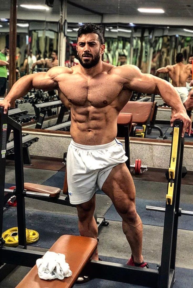 the image shows a muscular bodybuilder posing in a gym. he is shirtless, showcasing his highly developed muscles, and wearing white shorts. he is standing near a weightlifting bench and rack. the gym is visible in the background, with other gym equipment and mirrors. the bodybuilder's physique is the clear focus, highlighting his significant muscle mass and definition.  the overall lighting and composition emphasize the subject's muscular form.