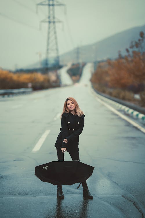a young woman in a black doublebreasted coat and black boots stands on a wet road holding a closed black umbrella.  the background is slightly blurred but shows a highway stretching into the distance under a gray sky.  there are power lines and some autumnal vegetation in the far background. the overall mood is moody and atmospheric.
