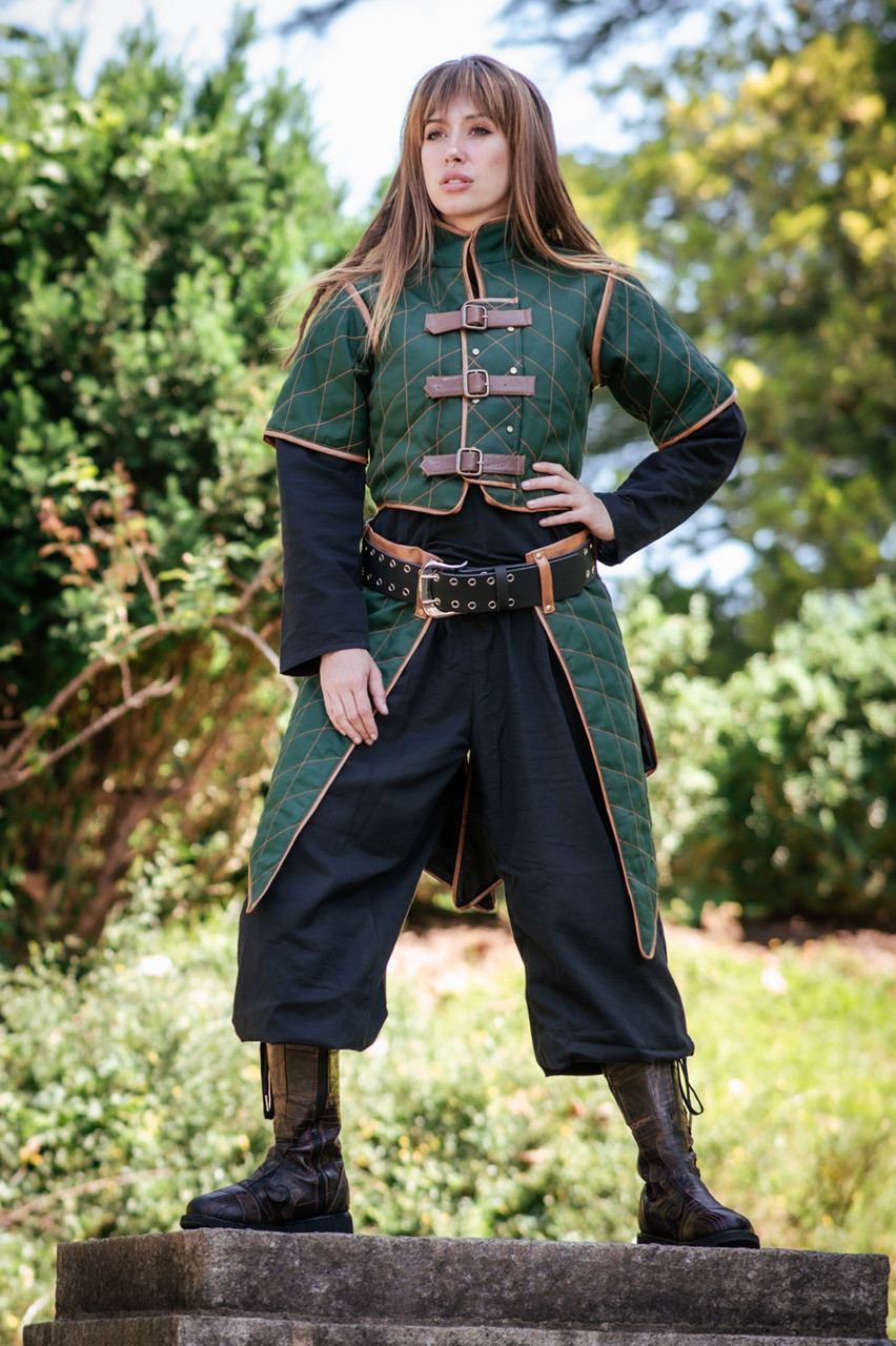 a woman in medieval clothing stands on stone steps. she is wearing a quilted green jacket with brown trim and leather straps, a black shirt, black pants that end below the knee, and black boots. she has a black belt with silver studs around her waist. there is greenery in the background.