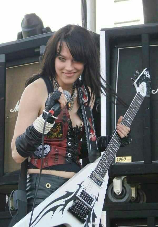 a young woman with long black hair is performing on stage with a white and black guitar. she is wearing a black leather jacket, a red corset, and black pants. she is holding a microphone and singing into it. there is a black guitar amp behind her.