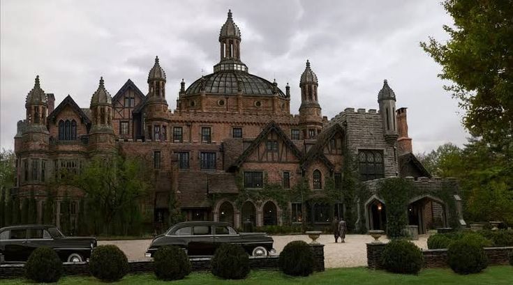 he image shows a large, imposing mansion that resembles a castle. it's made of brick and stone, with gothic and tudor architectural features, including towers, turrets, and arched windows. the building is covered in ivy in places, adding to its aged and somewhat mysterious appearance.  two vintage cars are parked in front of the house. the sky is overcast, and the overall mood of the image is dark and somewhat gloomy. the setting appears to be a large estate with landscaping around the house and driveway.