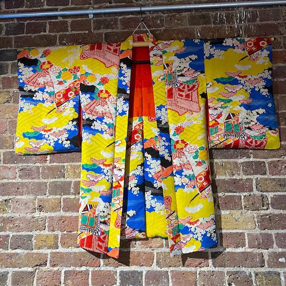 a vibrant, ornate kimono hangs on a hanger against a brick wall. the kimono features a bright yellow base with blue, white, and red patterns, suggesting traditional japanese designs. the interior of the kimono has alternating panels of red, orange, and blue. the brick wall provides a textured backdrop.