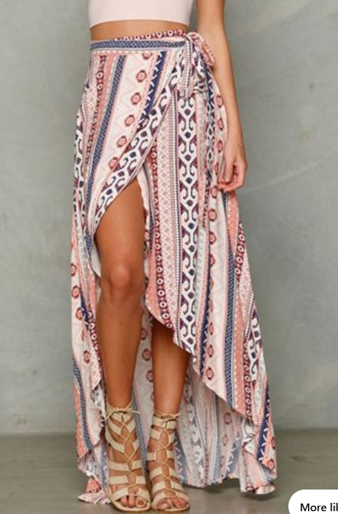 a woman is modeling a long, flowy wrap skirt with a colorful bohemian print. the skirt has a high slit up the leg and is paired with a simple pink top and strappy sandals.