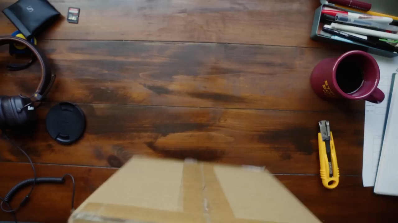 a cardboard box is being placed on a wooden desk. the image is taken from a firstperson perspective, as if the viewer is receiving the package.  the desk also has headphones, a camera lens cap, a coffee mug, and stationery.