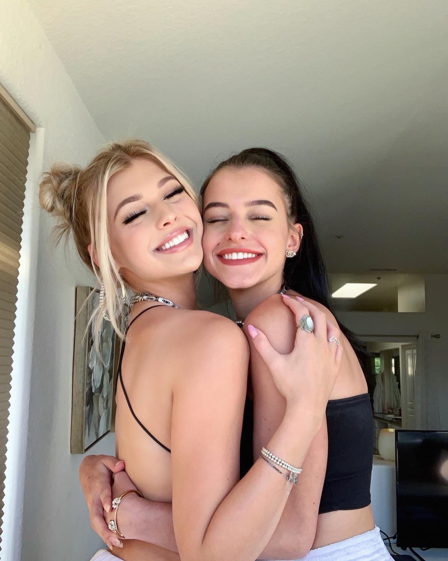 two young women are embracing each other and smiling. the woman on the left has blonde hair styled in a messy bun and is wearing a black tank top, white shorts, and a silver necklace and bracelet. the woman on the right has brown hair pulled back in a ponytail and is wearing a black tank top, white shorts, and a bracelet with charms and rings on her fingers. they both have their eyes closed and are smiling widely. the background appears to be an interior setting with a door and a window.