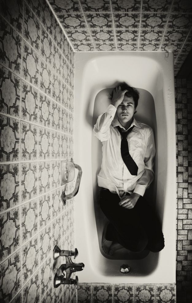 a black and white photograph of a man sitting in a bathtub. he is wearing a white shirt and a dark tie. his head is in his hands and his expression is one of sadness or despair. the bathroom is tiled with a vintagelooking pattern. the overall mood is dark and moody.