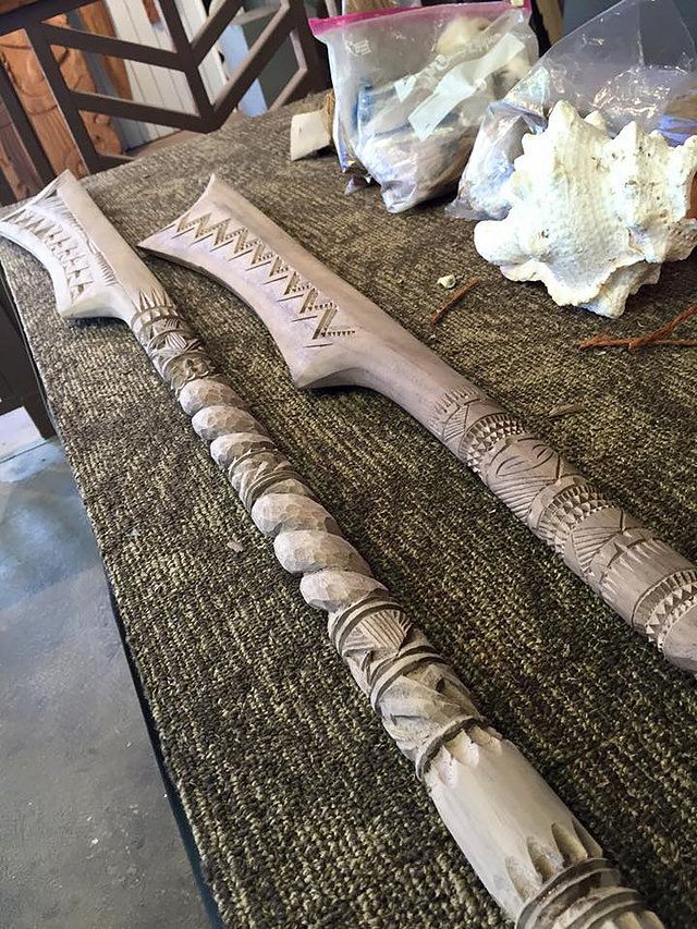 a highangle shot shows two handcarved wooden weapons on a textured surface, beside plastic bags and a shell. the weapons are decorated with intricate tribal designs and have different shaft designs, one with a spiral pattern.