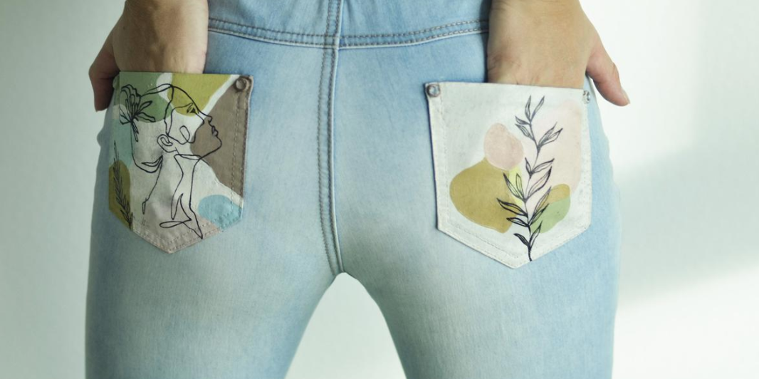 a rear view of a woman modeling a pair of light blue jeans. the jeans feature unique back pockets with an artistic design of a woman's profile and floral elements in pastel colors. the woman has her hands in her back pockets, showcasing the design.