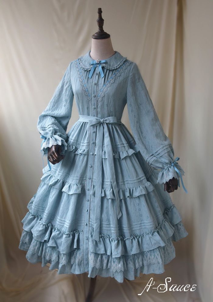 a light blue lolita fashion dress is shown on a mannequin. the dress has long sleeves, a lace collar, and a ruffled skirt. it is tied at the waist with a matching belt. the dress is made of a light, airy fabric and has a vintage feel.