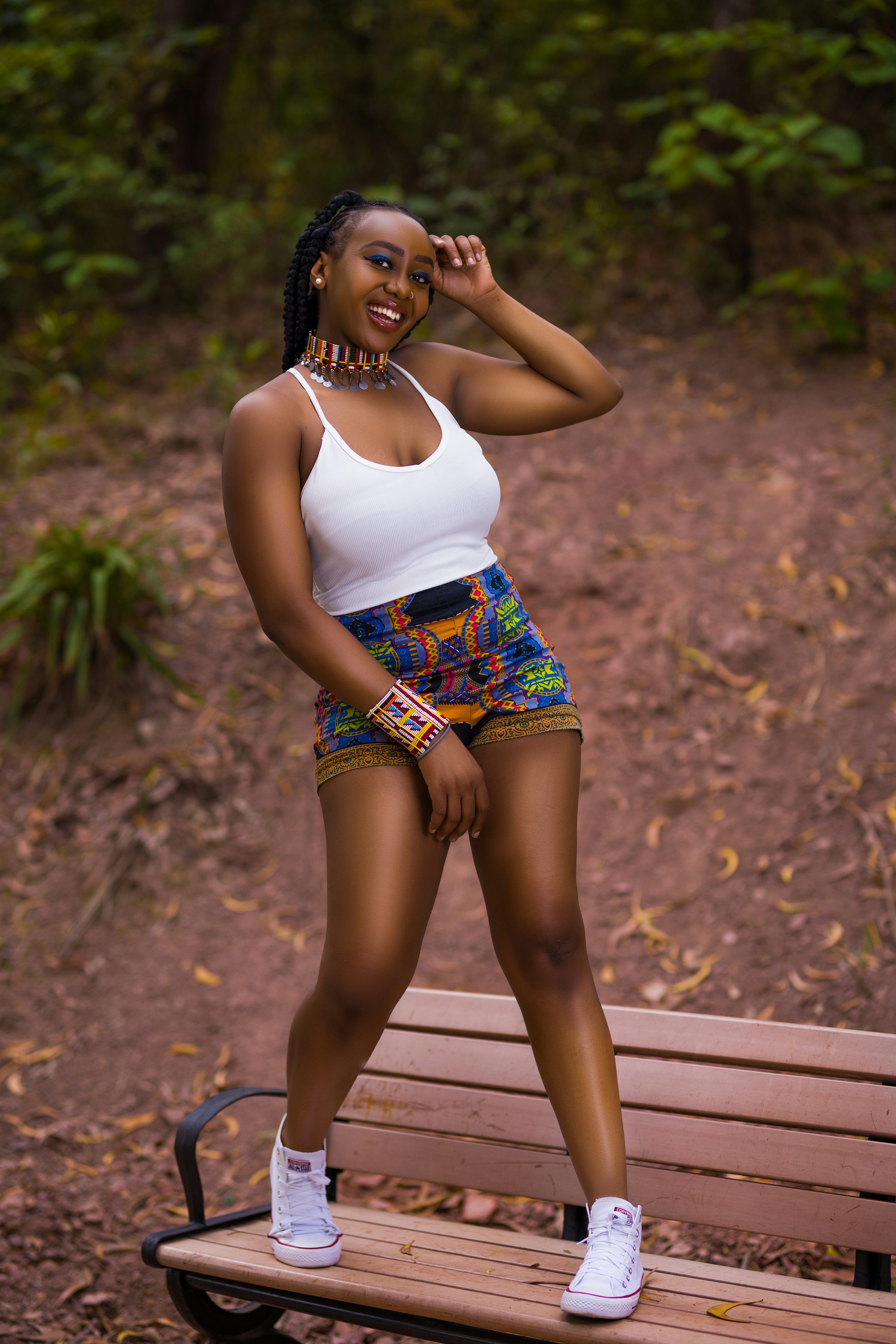 a young, darkskinned african woman with braids smiles confidently as she sits on a park bench. she's wearing a white tank top, vibrant africanprint shorts, beaded jewelry, and white converse sneakers. her posture is relaxed, and her expression is joyful. the background is a natural, outdoor setting with trees and earthtoned ground. the overall image conveys a sense of youthfulness, confidence, and cultural pride.