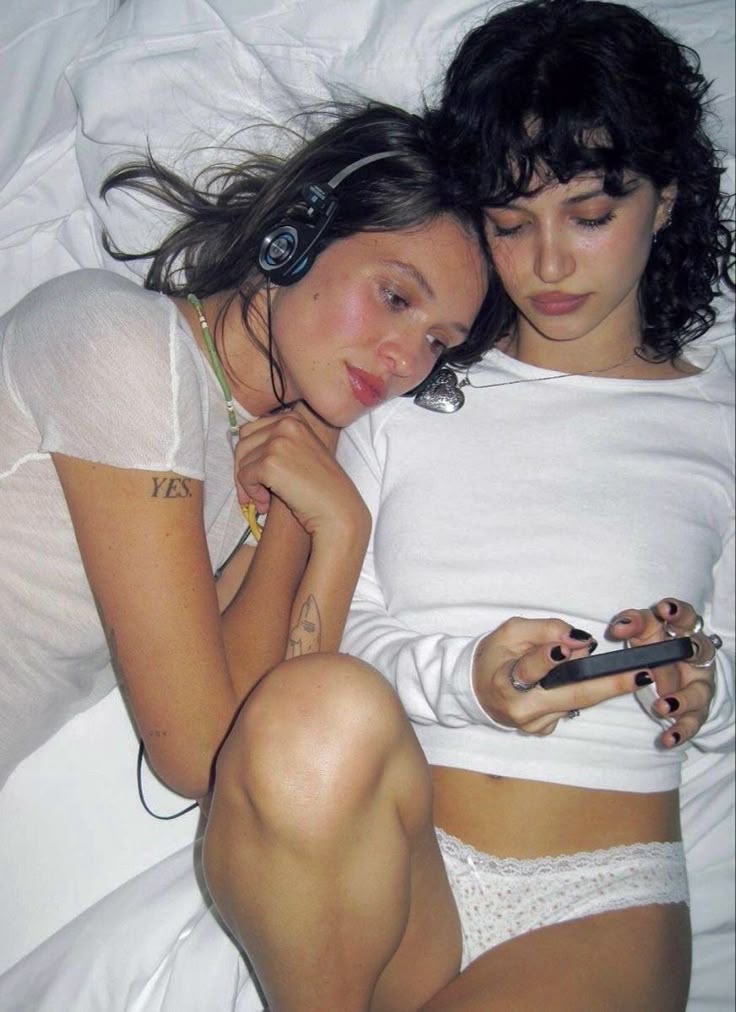 two women are lying on a white bed. the woman on the left is wearing headphones and a white shirt, and she has a tattoo on her arm. the woman on the right is wearing a white longsleeve shirt and white lace underwear, and she is holding a smartphone. both women are wearing jewelry around their necks.