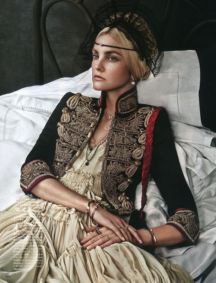 the image is a highfashion photograph showcasing a blonde model wearing an elaborate ensemble. she is seated on a bed, dressed in a flowing, light beige gown beneath a dark, richly embroidered jacket with militarystyle details.  the jacket features gold and black embellishments and red accents. her head is adorned with an intricate black lace headpiece. she wears numerous rings and bracelets, adding to the opulent feel. the overall style is romantic and vintageinspired, evoking a sense of oldworld glamour. the setting is a luxurious bedroom, adding to the feeling of elegance and sophistication. the lighting and styling contribute to a moody and atmospheric scene.