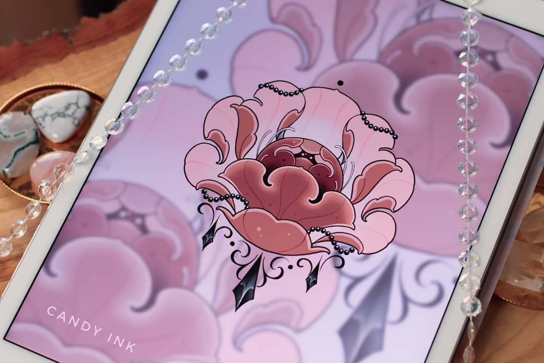 a digital drawing of a pink flower with black accents in a neotraditional tattoo style is displayed on a tablet.  the background of the drawing is pink and purple clouds.  surrounding the tablet are quartz crystals and a strand of iridescent beads.  the watermark "candy ink" is visible on the bottom lefthand side of the image.