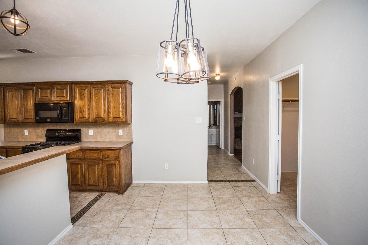 a kitchen with wooden cabinets and countertops, a kitchen island, and a lighting fixture. a hallway leads to a bathroom and a closet. the floor is tiled.