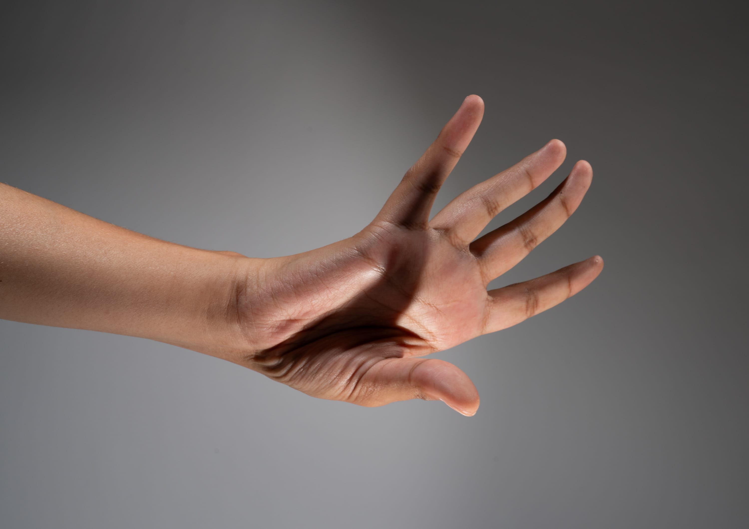 a closeup side view of a hand extended with fingers spread apart, dramatically lit by a spotlight against a grey background. the contrast of light and shadow highlights the texture and contours of the skin.