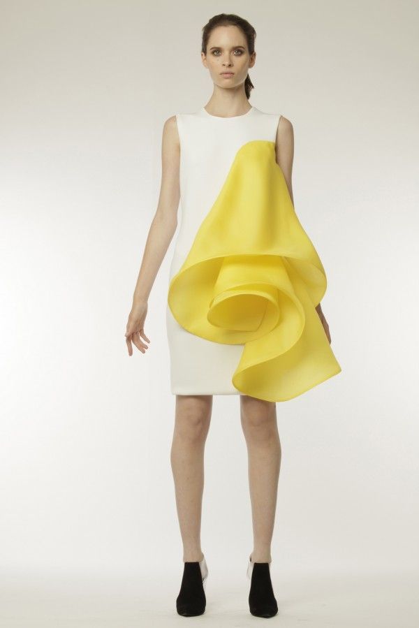 a woman in a white dress with a yellow, voluminous, roselike accent on one side stands with a straight face. the dress is sleeveless and fits closely to the body. she wears her hair down with a center part and has natural makeup on. she wears black heels with white accents on the heel. the backdrop is a simple white background.