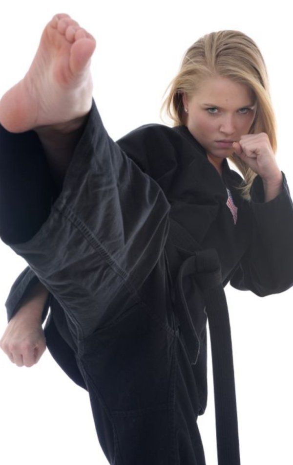 a young woman dressed in a black karate uniform, with a black belt tied around her waist, throws a powerful kick with her right leg, as she looks intently toward the camera. she has shoulder length blonde hair. her left hand is clenched into a fist, and her left leg is bent at the knee, as if she's preparing for a second move.