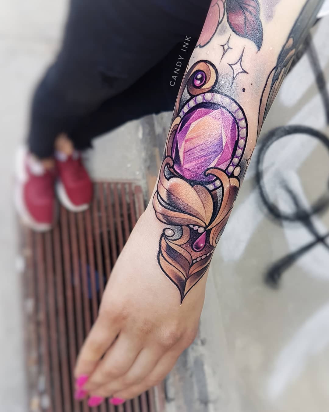 a close up image of a  woman's forearm. she has a neotraditional tattoo of a purple crystal on her inner arm. the crystal is surrounded by ornate gold and brown decorations. there is a small heartshaped droplet hanging from the bottom.