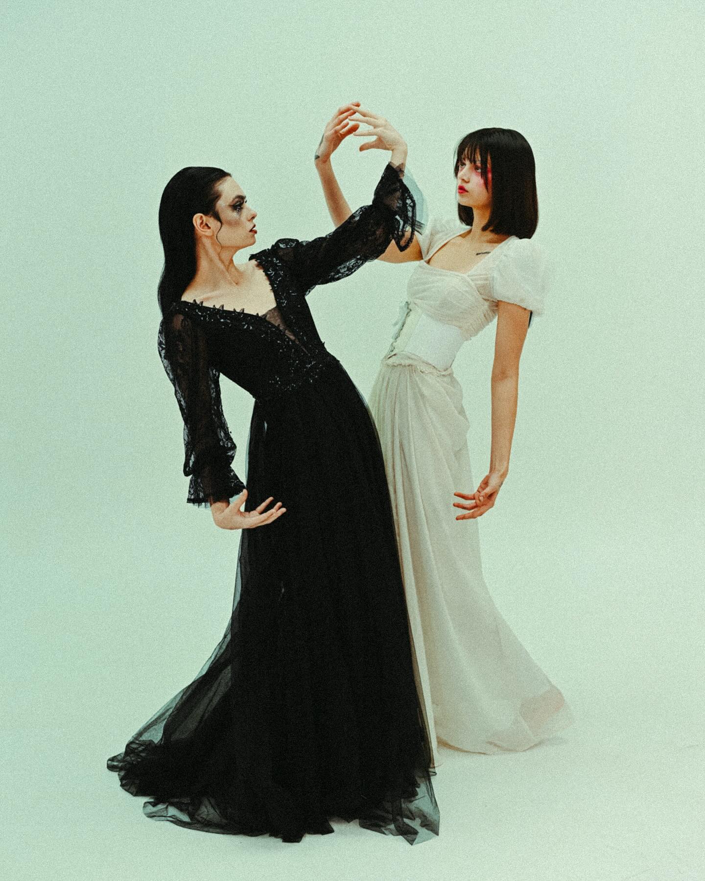 two women are featured in a studio shot. one woman is wearing a long, black tulle dress with lace details, and the other is wearing a long, white chiffon dress with a corsetlike bodice. the women are posed gracefully, their bodies interacting. the image has a dark and light contrast, with the dark dress representing gothic style and the white dress representing lightness and purity. the overall aesthetic of the image is elegant, artistic, and stylish.