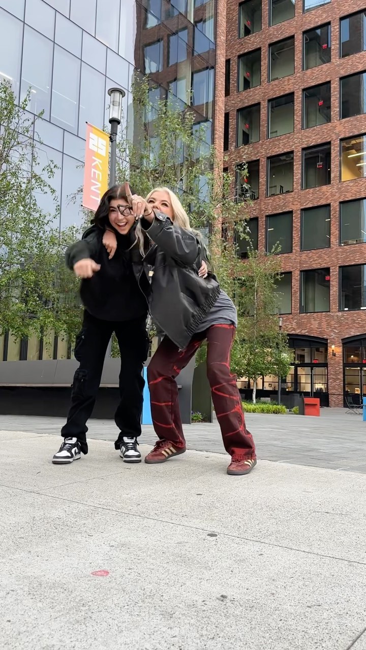 two women pose outside a modern building, one wearing black clothing and sunglasses, the other in a leather jacket and red pants. they are standing on a sidewalk with trees and urban architecture in the background.