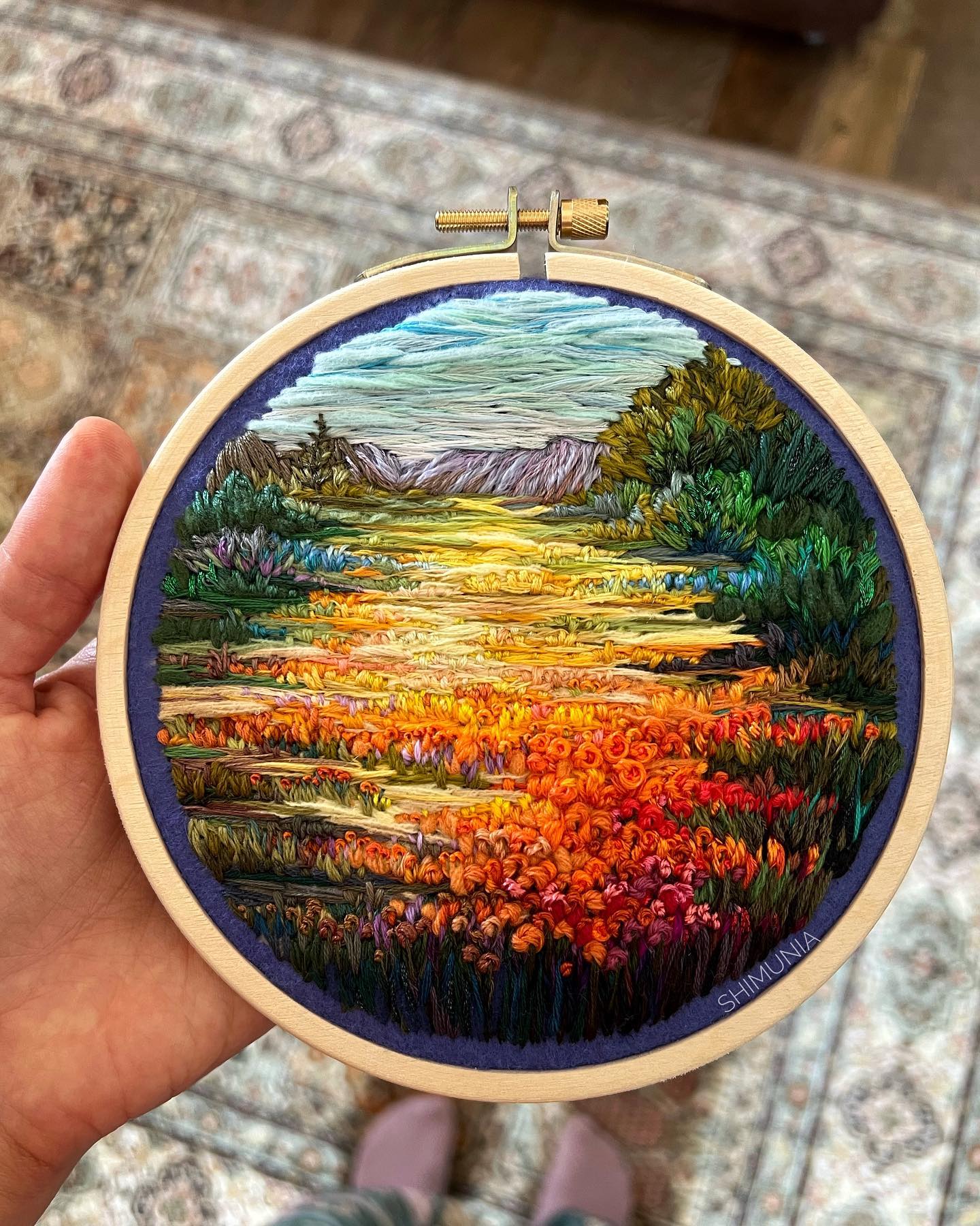 a person is holding up a finished embroidery hoop. the embroidery is a landscape scene of a sunset over a field of orange flowers with mountains in the distance. it is signed "shimomnia" on the bottom right of the hoop.