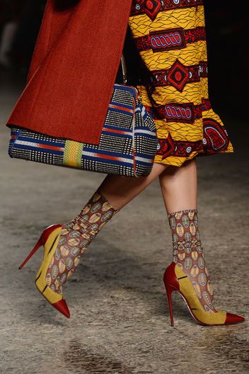 a closeup shot of a fashion model's legs and lower body, showcasing a striking outfit. the model is wearing a vibrant yellow and red african print skirt, a rich red coat, and eyecatching patterned socks. her shoes are highheeled pumps with a unique twotoned design in yellow and red. the model carries a multicolored handbag that complements the overall aesthetic. the image is taken on a runway or similar setting, suggestive of a fashion show or editorial shoot. the overall style is bold, chic, and incorporates a mix of textures and patterns. the focus is on the details of the outfit, specifically the contrasting colors and patterned elements, which make the overall look very visually appealing.