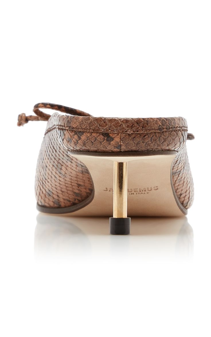 the image shows a rear view of a pair of jacquemus women's mules. the shoes are lowheeled, featuring a unique gold metal heel. they are made of brown snakeskin leather with a knotted detail at the top. the "jacquemus" brand is subtly visible on the sole.  the background is plain white, allowing the shoes to be the focus. the image is a highquality product shot showcasing the shoe's detail.