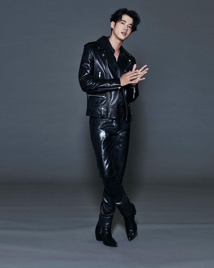 a full body shot of a stylish asian man in a black leather jacket and matching pants. he is wearing black shoes and has his hands clasped. the image is against a gray background.