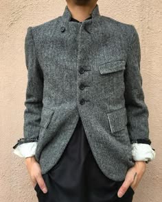 a man is wearing a grey tweed jacket with a standup collar and buttons. the jacket has pockets on the front and the sleeves. he is standing and looking at the camera.