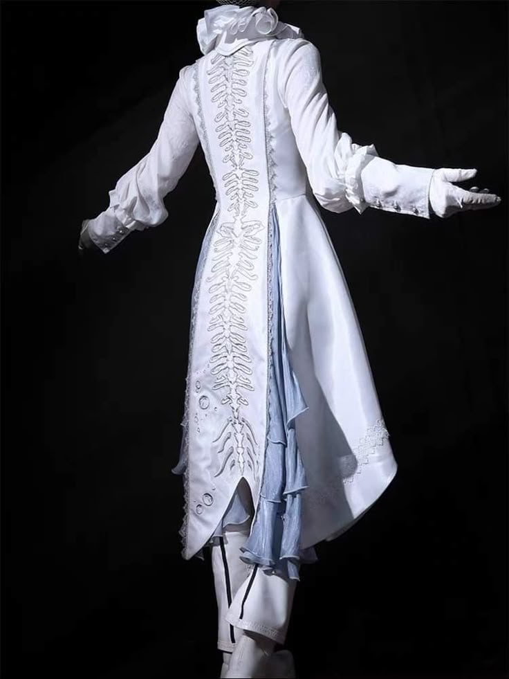 a person is facing away from the viewer, wearing a long white coat with intricate white embroidery along the back. the coat has a high, ruffled collar and long sleeves with ruffles at the cuffs. a blue fabric panel peeks out from underneath the coat. the person is also wearing white pants. the background is dark and unclear.