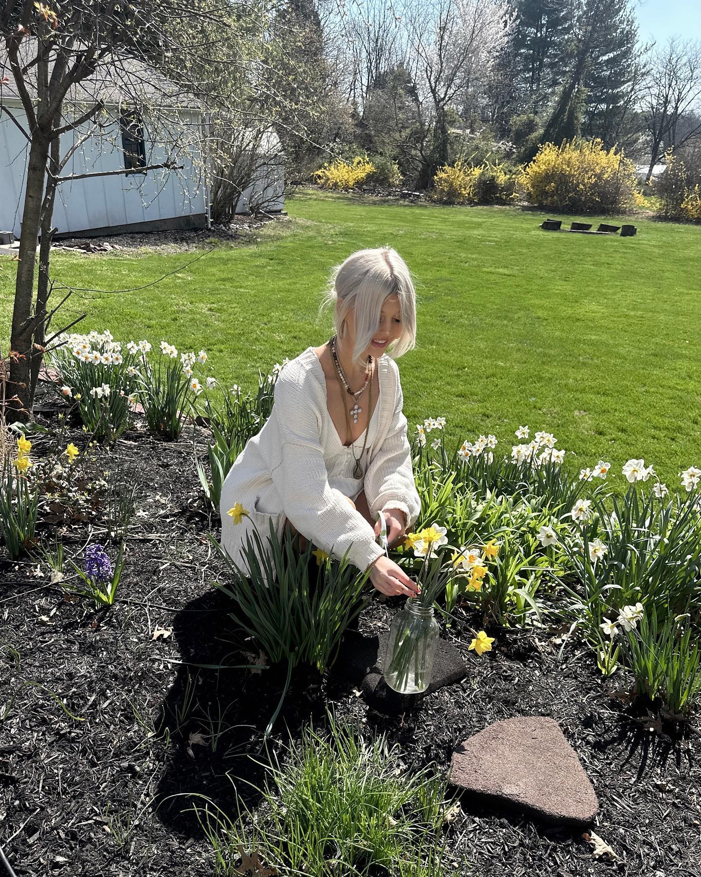 a young woman with blonde hair is kneeling in a garden bed full of daffodils. she is wearing a white sweater and has several necklaces on. she is arranging the daffodils into a glass jar. the garden bed is located in a yard with a green lawn and trees. in the background is a house.
