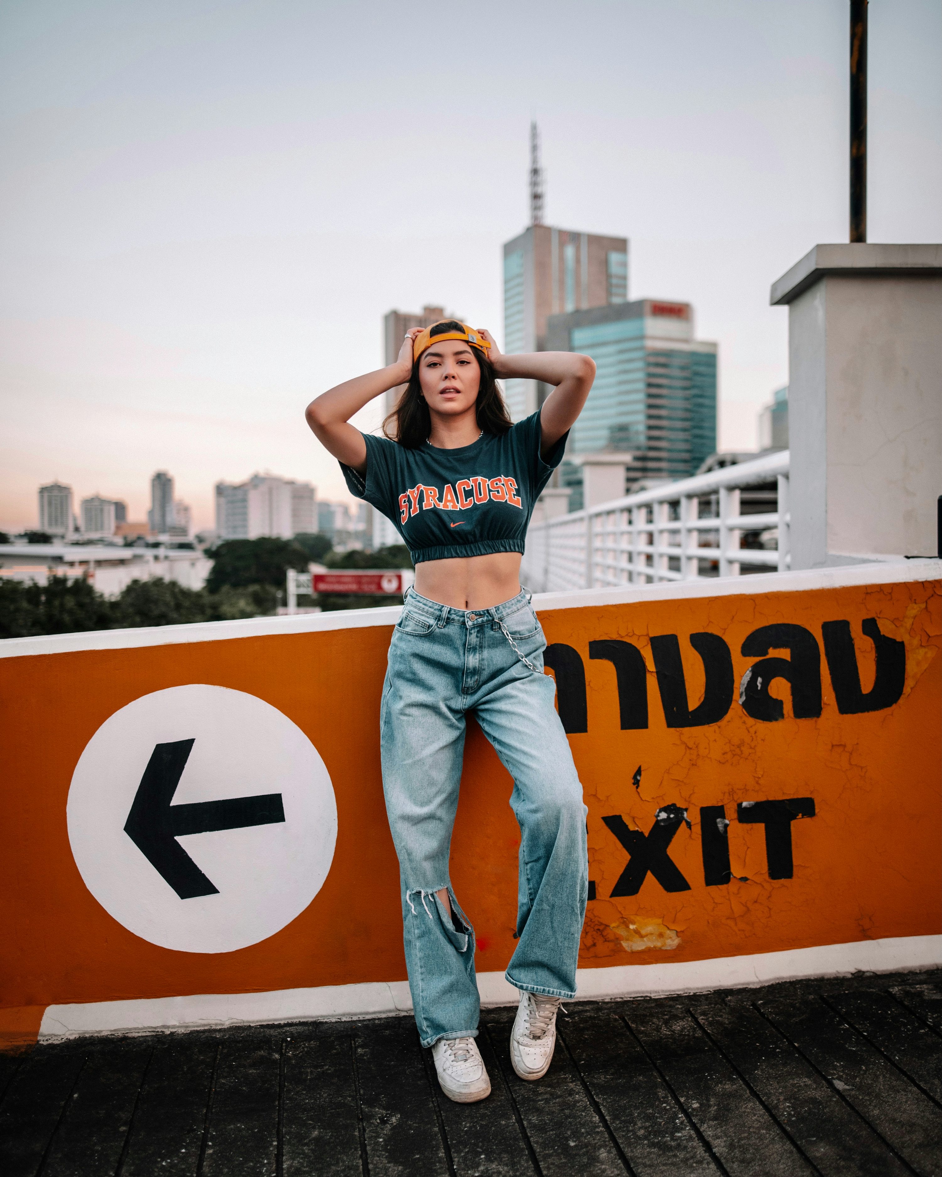 a young woman in a stylish outfit poses against an orange wall with a directional sign. she wears a syracuse crop top, baggy light blue jeans with rips at the knees, a yellow cap, and white sneakers. the backdrop is a city skyline with skyscrapers at sunset. the overall mood is trendy and casual.