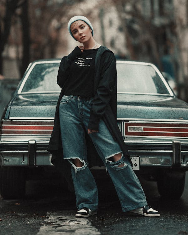 a stylish young woman wearing ripped jeans, a black long cardigan, and a light blue beanie is standing next to a vintage car.  she is posing for a photo, looking towards the camera. the setting appears to be an urban street. the overall aesthetic is cool, casual, and trendy. the image has a slightly moody and atmospheric tone.