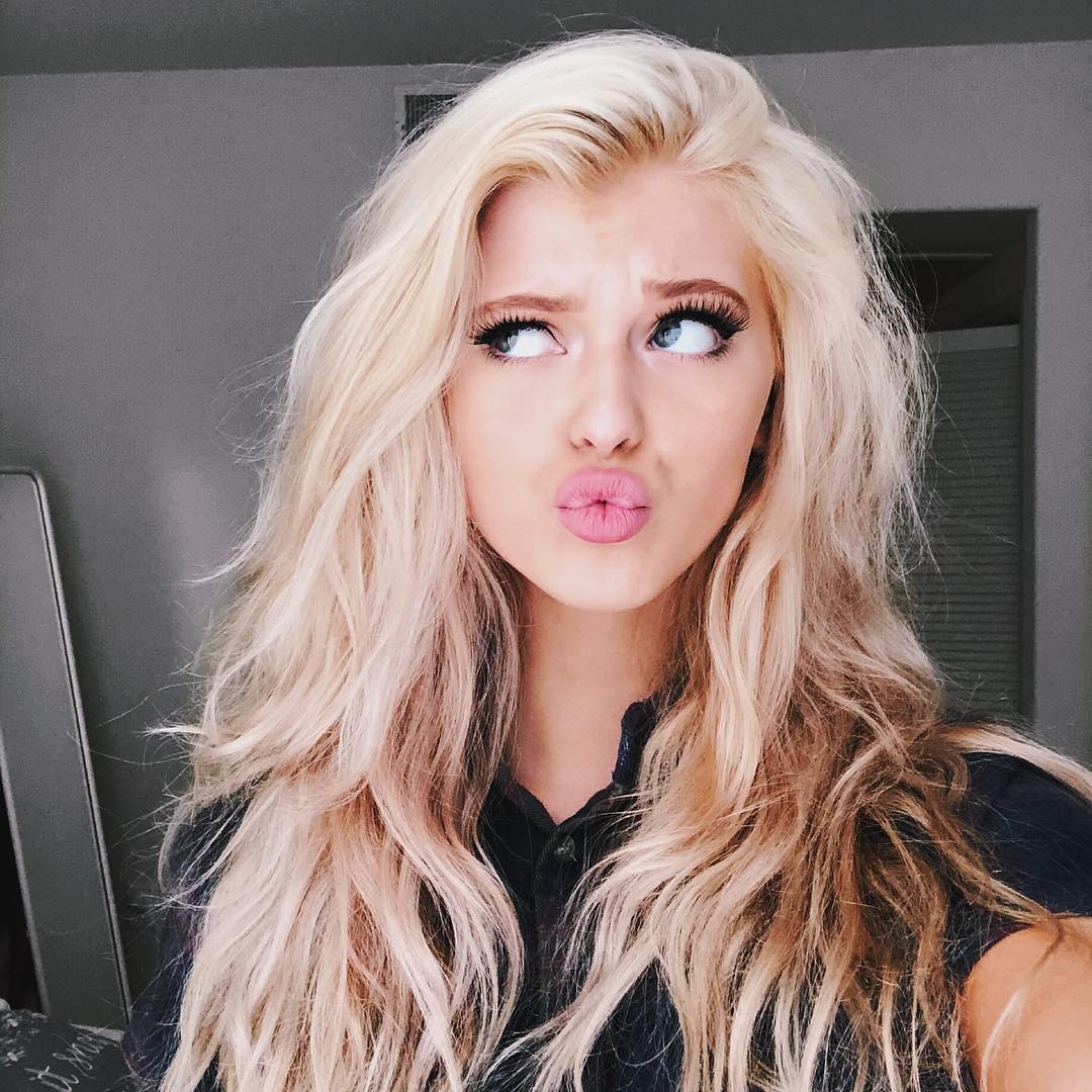 a woman with long, blonde hair and blue eyes is pouting in a selfie. she is wearing makeup and has long eyelashes. she is wearing a black shirt and is indoors.