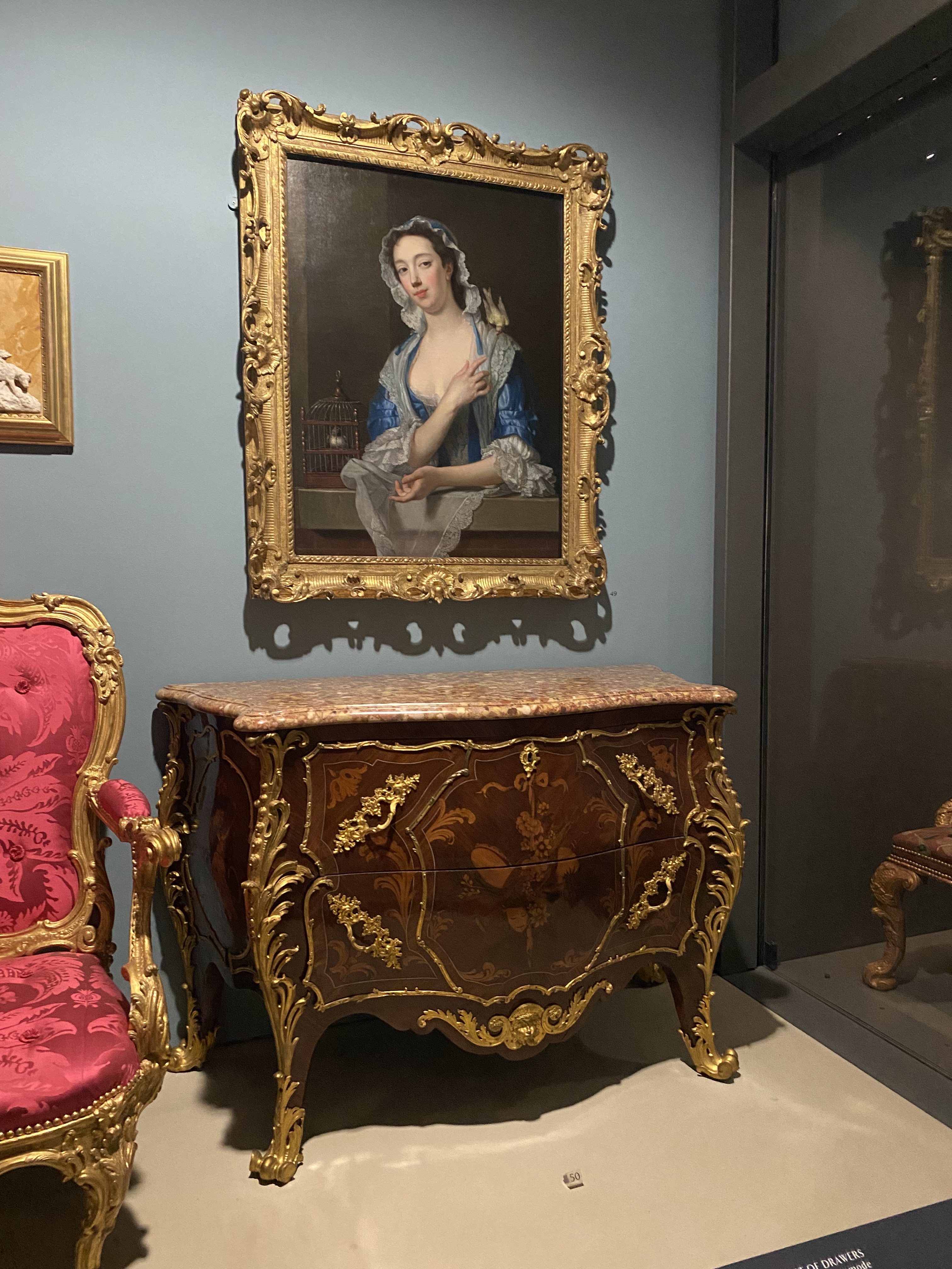 a photo of a museum display with a painting and furniture. the painting is a portrait of a woman in a blue dress with a white lace collar and a small bird on her shoulder. the painting is in a large, ornate gold frame. below the painting is a chest of drawers with a marble top. the chest of drawers is also ornate with gold accents and brown wood paneling. to the left of the chest of drawers is a red chair with gold accents. to the right of the dresser is a glass case. the background is a blue wall. the overall style is baroque/rococo.