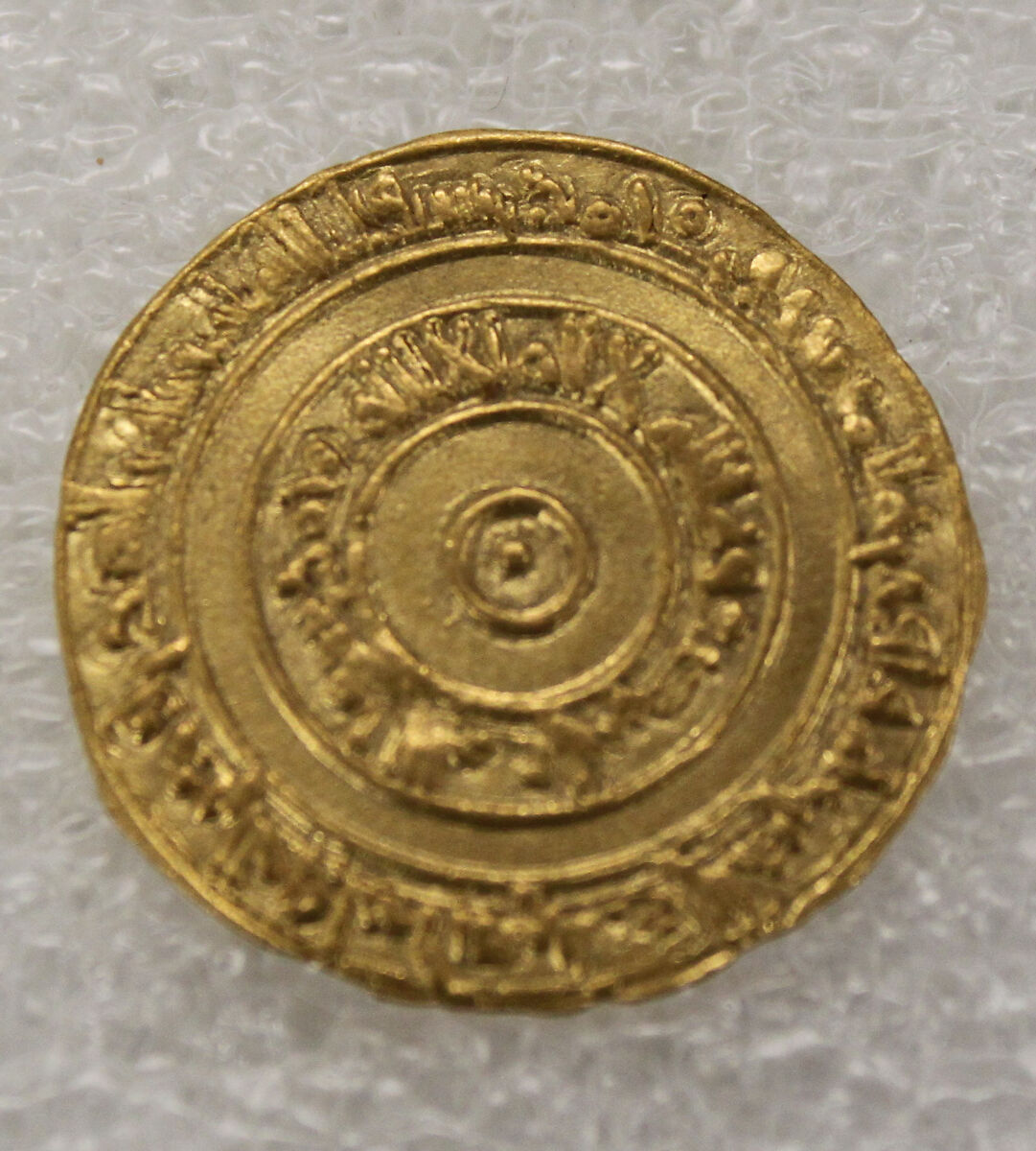 the image shows a golden, circular coin with inscriptions. it appears to be an ancient artifact, likely a numismatic item of historical significance. the coin has a central design with concentric circles and inscriptions around the edges. it is presumably made of gold and shows signs of wear and age.