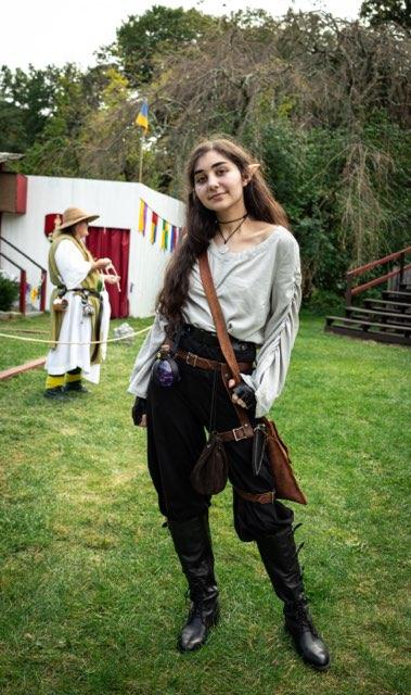 a young woman dressed in an elf costume stands on a grassy field at a renaissance fair. she has long brown hair, pointed ears, and is wearing a grey shirt, black pants, and tall leather boots. she has a leather belt with a pouch and bag attached to it. in the background, there's a man dressed in a medieval costume and a building with decorations. the scene is set outdoors in a field with some trees visible.