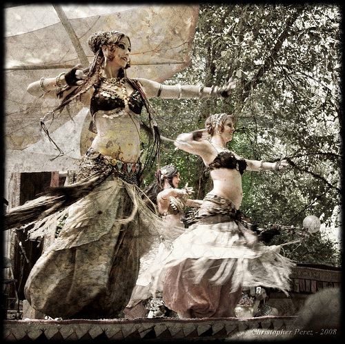 two women in ornate costumes perform a belly dance in an outdoor park setting, surrounded by trees and greenery.  the dancers are in full motion, their bodies moving fluidly and gracefully as they show off their skills and the beauty of their costumes.  one of the women is wearing a long flowing skirt with fringe, while the other is wearing a shorter skirt with a more layered look.  both women are wearing ornate headpieces and jewelry, and their makeup is dramatic and expressive. the scene is full of energy and movement, and the dancers are clearly enjoying themselves.  the setting is picturesque, with the lush greenery providing a beautiful backdrop for the performance.  the image captures the beauty and passion of belly dancing and the joy of celebrating cultural traditions.