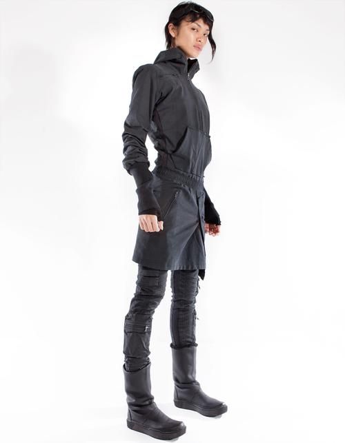 a full studio shot features a woman standing in black clothing and boots, evoking a cyberpunk aesthetic. she is wearing goggles and a coatlike garment.
