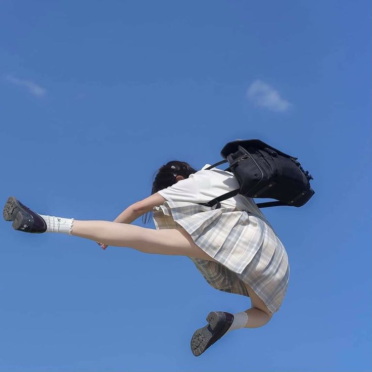 a woman, dressed in a school uniform with a pleated plaid skirt, white socks, and oxford shoes, is jumping in midair against a clear blue sky. she has a black backpack on. her legs are extended, and she appears to be in motion. she has a hair clip in her hair. the picture seems to be taken during the day in the summer.