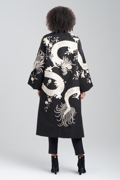 a woman stands facing away from the viewer, showcasing the back of a long, black coat adorned with intricate embroidery. the focal point of the coat is a large, stylized dragon motif that winds across the back, complemented by floral embroidery. she is wearing black pants and ankle boots, completing her outfit. her curly hair adds texture to her silhouette. the background is a plain, light gray, emphasizing the details of her attire.