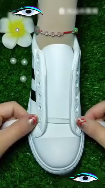 a closeup shot shows a person's hands lacing up a white sneaker that is resting on artificial turf. a white flower, a few pearls and an ankle bracelet are in the shot.