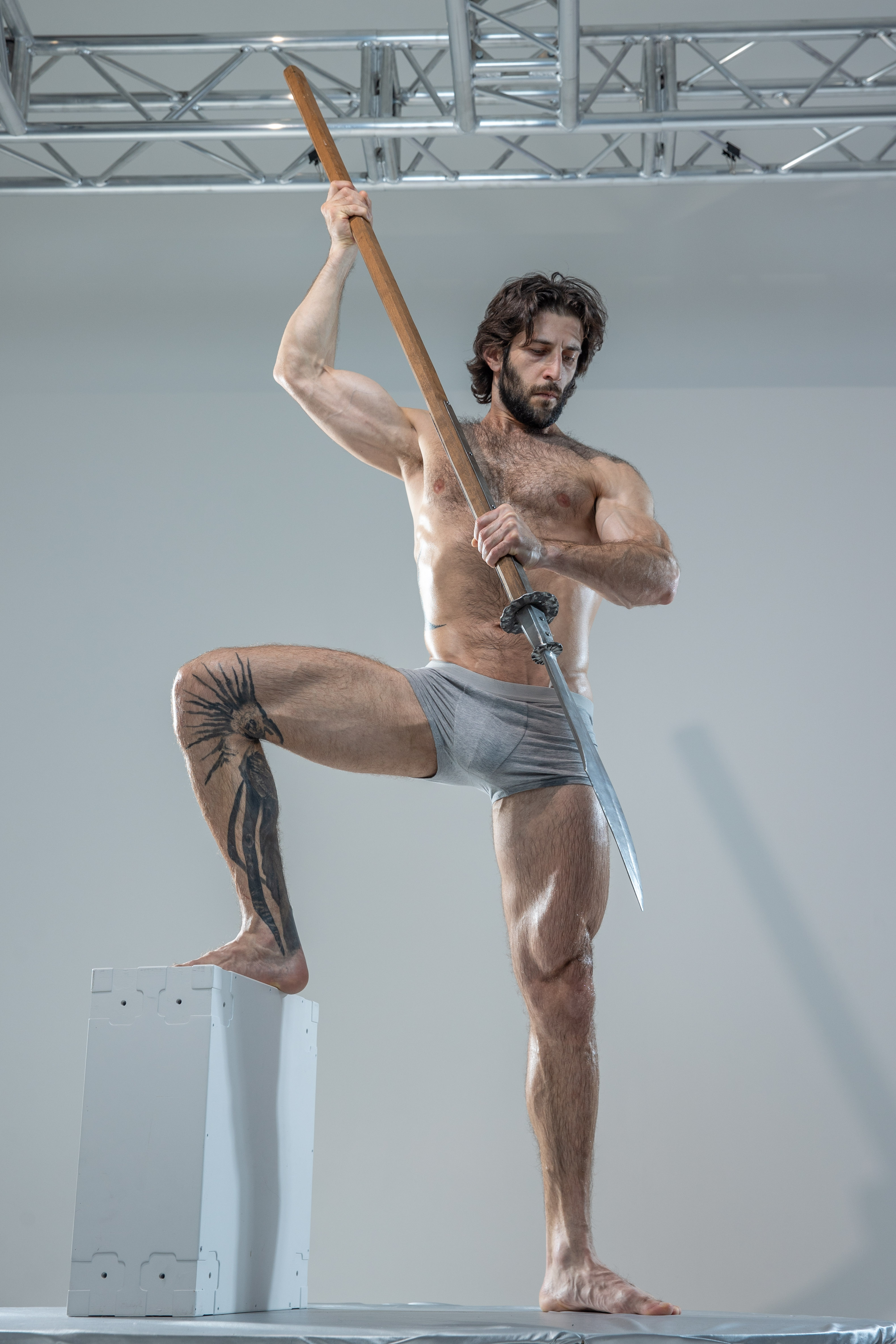 a muscular man with a beard and tattoos stands on a white platform in a bright studio. he holds a spear aloft in a powerful pose, showcasing his physique. he wears only grey underwear, allowing for a clear view of his musculature. the background is a simple grey wall, with metal scaffolding visible at the top. the overall image evokes a sense of strength, power, and masculinity.