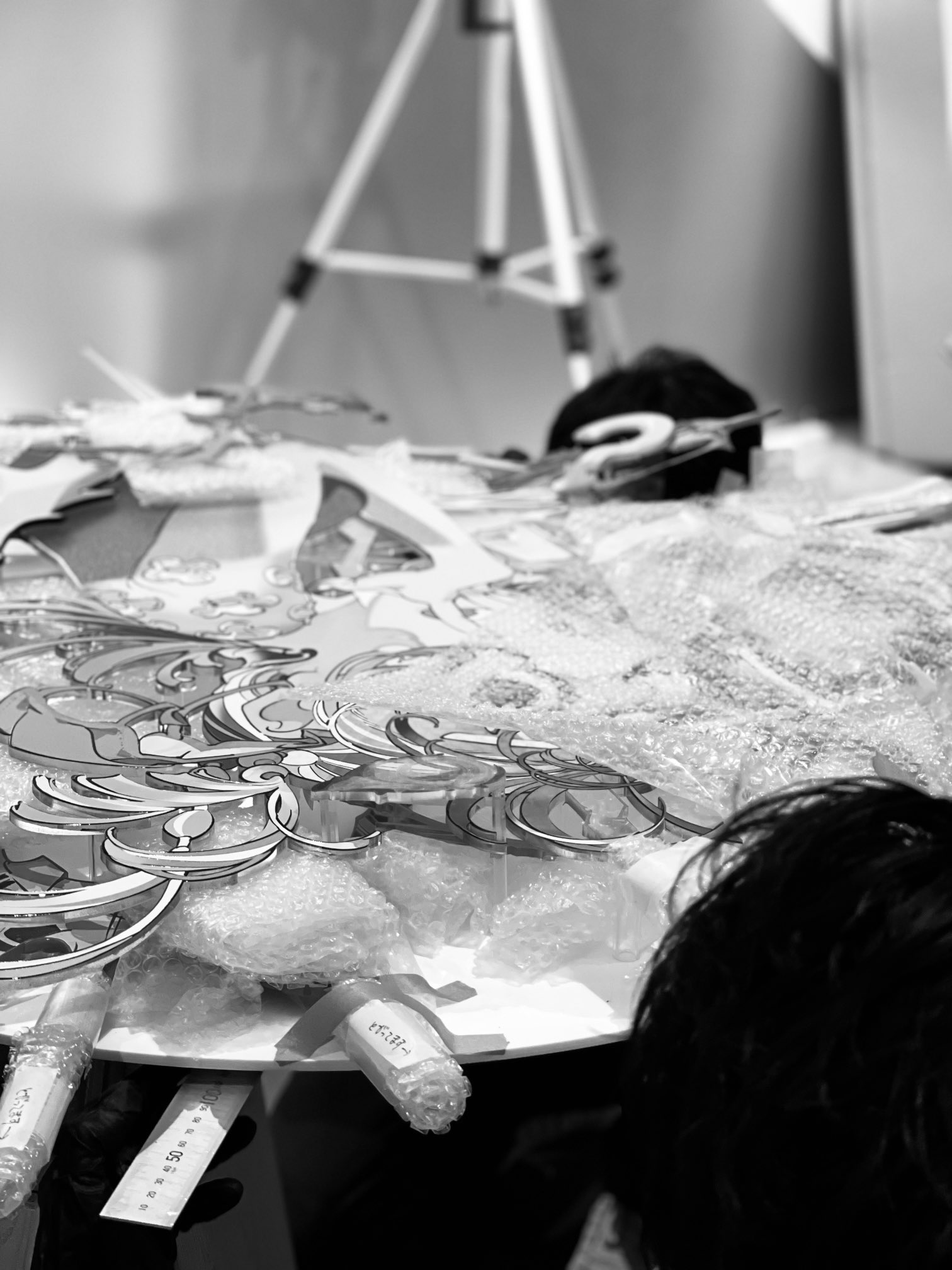 a black and white image showing the top of a round table where multiple people are assembling what looks to be a mixed media artwork. the artwork has intricate designs and is layered upon what looks like foam board and covered in bubble wrap. the people assembling the art are hidden behind the piece and only the tops of their heads are visible. the room is bright but the photo is dim, giving the image an overall grayscale palette.