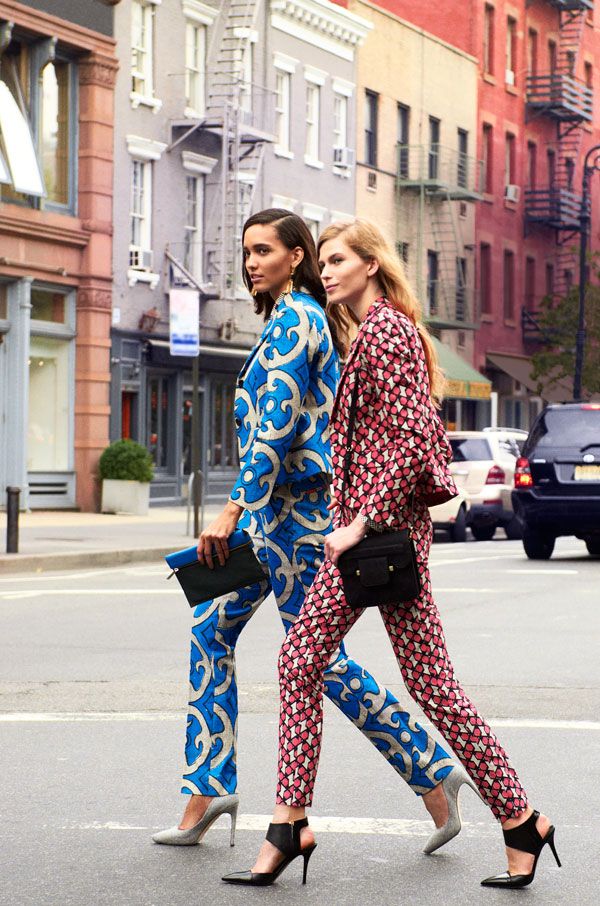 two women in stylish patterned pantsuits are walking across a crosswalk in a new york city street. one woman wears a blue pantsuit with a bold, abstract print, while the other woman wears a pink pantsuit with a smaller, geometric print. both women are wearing high heels and carrying handbags. the background features brick buildings and parked cars. the image showcases a modern, fashionable look incorporating bold prints and tailored silhouettes.