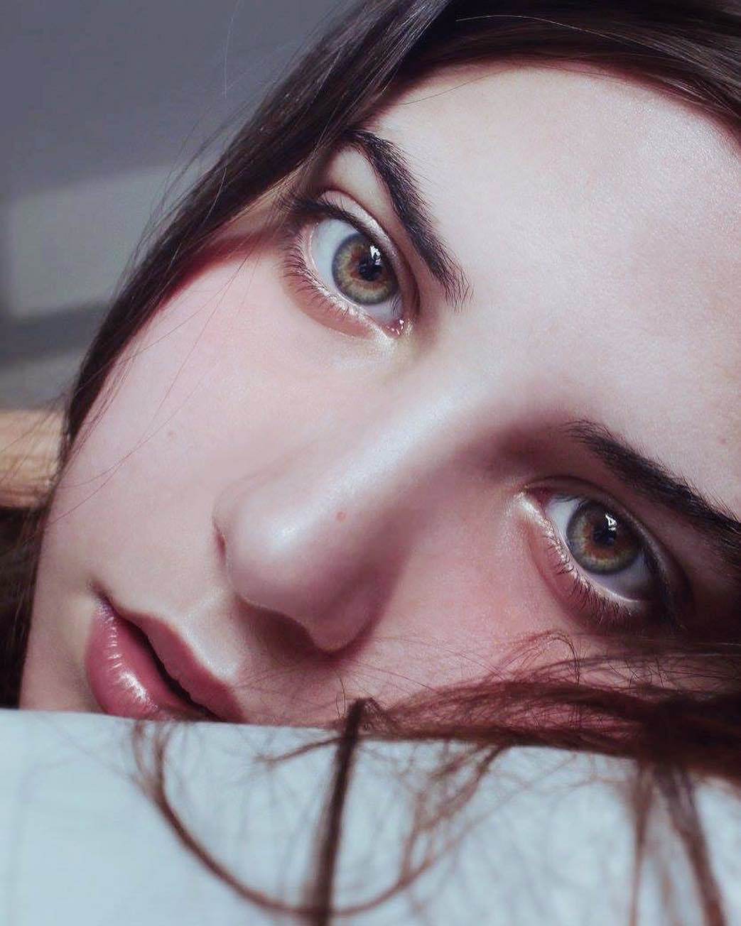 a closeup portrait of a woman's face, focusing on her eyes, nose, and lips. she has fair skin, dark hair, and light brown eyes with dark eyebrows. the image is slightly angled, and part of her hair rests on a white surface, possibly a pillow. her lips have a natural, glossy appearance.